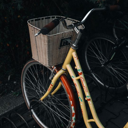 A yellow cruiser bike with cherry print detailing and a wicker basket parked alongside a dark bike in an outdoor setting.