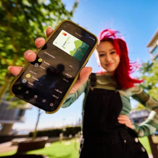 A young woman with bright red hair smiles and holds up a smartphone displaying the Clean Energy Credit Union app and debit card on a sunny campus.