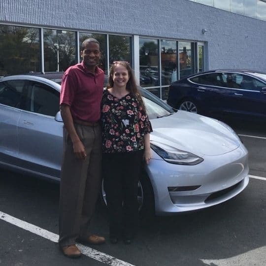 A man and woman smile together in front of their electric vehicle in a parking lot outside a building with large windows.