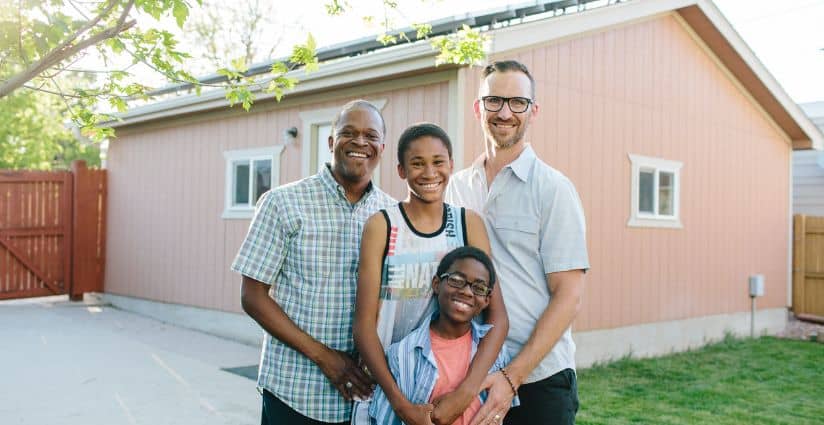 A family of four smiles together outside their home on a sunny day. Solar panels are visible on the roof behind them.
