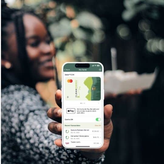 A person holds an iPhone displaying the Clean Energy Credit Union mobile app open to the My Cards screen, showing a green CECU debit Mastercard with an Add to Apple Wallet button and recent transactions listed below.