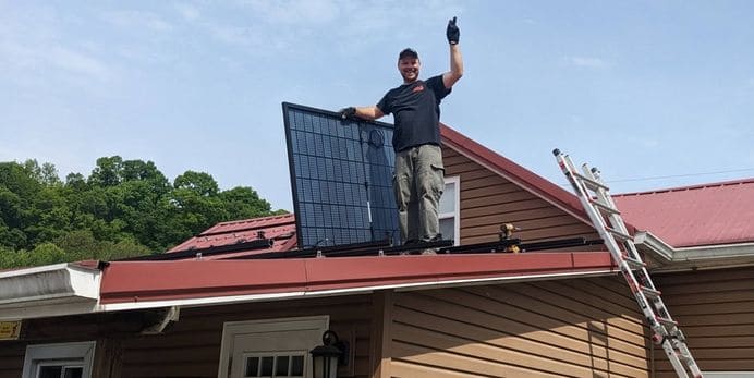 A photo of someone waving on a roof while holding a solar panel they are installing.