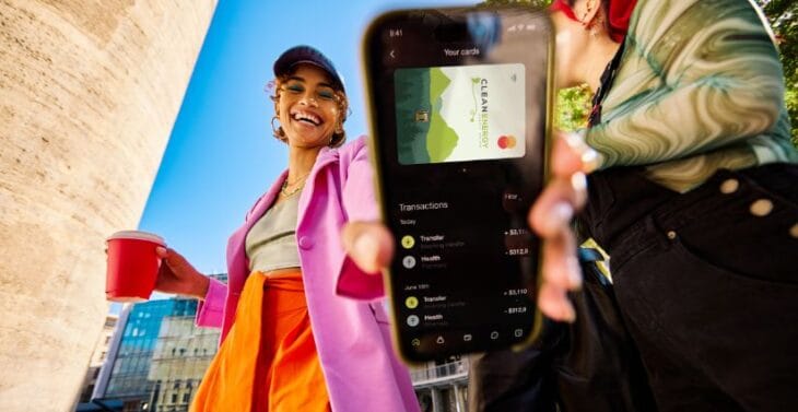 A young woman laughs outdoors on a sunny day while a friend holds up a smartphone displaying the Clean Energy Credit Union app and debit card.