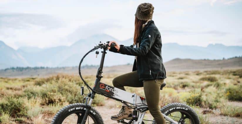 A woman in a jacket and knit hat sits on a fat tire folding electric bike overlooking a wide open mountain landscape.