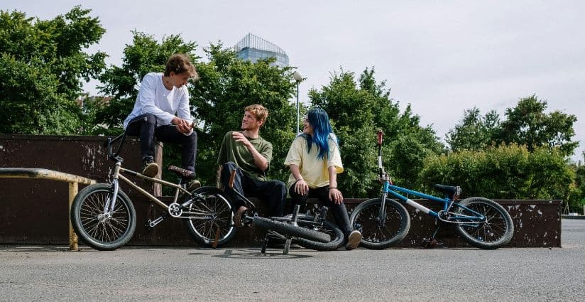 Three young people hang out and laugh together at an outdoor skate park with their BMX bikes resting nearby.